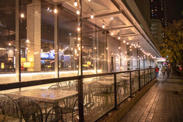 Empty restaurant patio tables and chairs under strings of light in the rain, glass railings, nighttime, nobody	