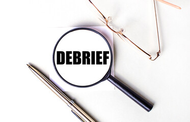 Golden pen and glasses, black magnifying glass with the text DEBRIEF on a light background.