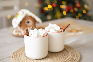 two white cup of cocoa with marshmallows and gingerbread house on table on background of Christmas tree