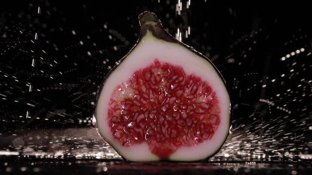 Ripe raw fig cut in half. The water is falling on the fig, drops of water are on the fruit. Black background. The fruit lays on the black table. Backlight.