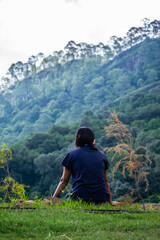 Naklejka premium A woman sitting and enjoying view of mountains and hills. Dense forest hill in the background creates beautiful landscape. Woman enjoying life in the mountains.Traveler on a remote place in the hills.