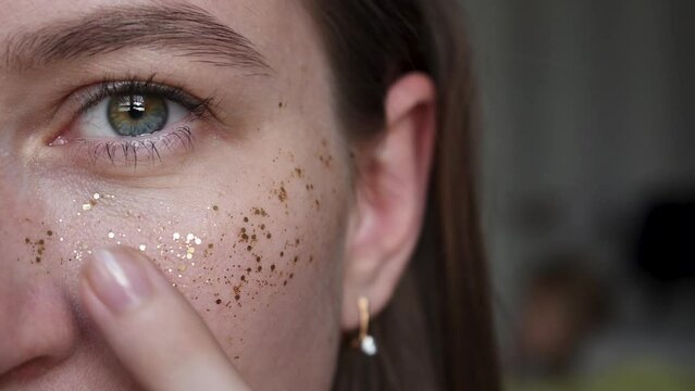 Close up of woman eye different colors look at the camera. puts glitter on face indoors. Partial heterochromia iridum. half of female face with golden sequins on cheekbone in the form of freckles. 