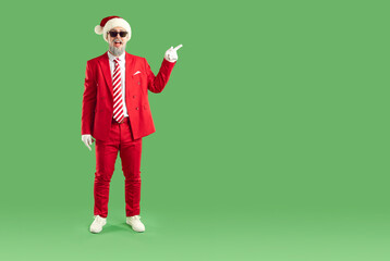Cheerful Santa with a smile on his face in sunglasses and a hat on a chroma key. An elderly bearded man in a red suit points with his index finger at an empty space for text on a green background.