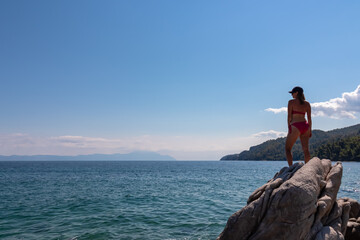 Woman in bikini standing on rock with panoramic view of holy Mount Athos from Fava sand beach, Vourvourou, Sithonia, Chalkidiki (Halkidiki), Greece, Europe. Summer vacation at Aegean Mediterranean Sea