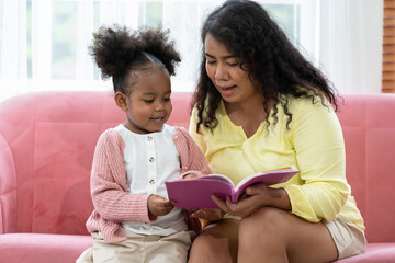 Mother and little daughter reading book together at home. African American child girl reading book with mom on sofa at home