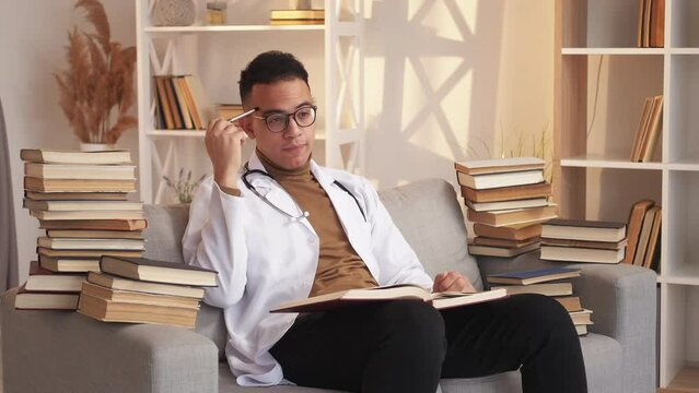 Learning medicine. Doctor training. Professional skill development. Pensive smart male intern studying thinking reading on couch with book pile at modern home interior.