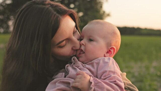 Happy baby sitting in mother's arms. Close-up shot of young good-looking woman kissing lovely daughter. Positive emotions. Horizontal view. Sunset on field in summertime. Footage of attractive