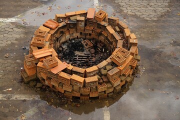Runde offene Feuerstelle aus rotbraunen Backsteinen auf nassem grauen Pflastersteinplatz mit Spiegelung bei Sonne am Nachmittag im Sommer