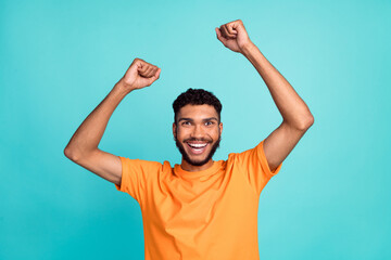 Photo of confident lucky guy wear orange t-shirt smiling rising fists isolated teal color background