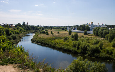 Fototapeta na wymiar Pastoral landscape - a beautiful bend of the Kamenka river among a green meadow with grass and a white stone monastery on the shore on a sunny summer day in Suzdal, Vladmir region, Russia