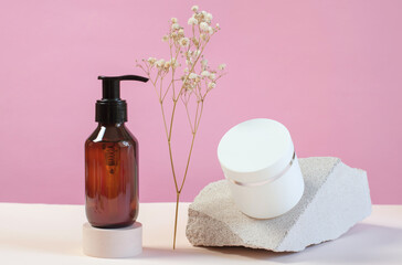 Fototapeta na wymiar A brown bottle with a dispenser with a gel and a white jar with a cream on a pink background. Skin care products on the background of white stones and gypsophila.