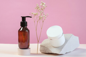 A brown bottle with a dispenser with a gel and a white jar with a cream on a pink background. Skin care products on the background of white stones and gypsophila.