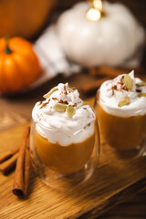 Pumpkin spice latte in glass mug with cinnamon, nutmeg, pumpkin seeds and whipped cream on brown texture wood. A glass of autumn coffee with pumpkin. A warming drink. Autumn concept.