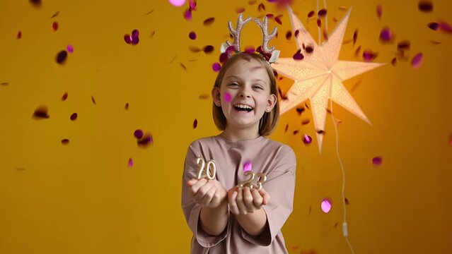 A happy girl with deer horns on her head celebrates the New Year, holds the numbers 2023 in her hands and laughs. Confetti fireworks on a yellow background.