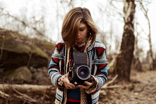 A beautiful young caucasian brunette woman standing on a forest path with a vintage camera in her hands taking a picture.