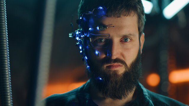 Brunette guy with dark beard wearing futuristic headset with one-eyed glasses, mic and ear piece looks at the camera with intense facial expressions. Neon lights in the background. Cyberpunk.