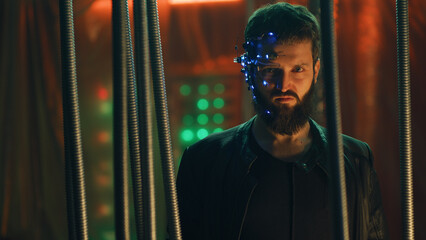 Young brunette guy in Cyberpunk style stands behind the black rods hanging. Wearing a futuristic...