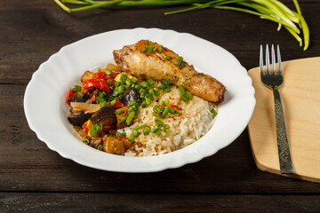 Rice with vegetables and chicken leg in a white plate on a handmade wooden board next to a fork on a wooden table.
