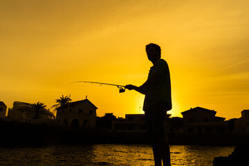 Angler in silhouette looking towards the sea before launching the fishing rod, behind a coastal town and the sea. orange sunset background,
