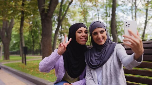 Happy muslim women talking selfie on smartphone in park, friends hanging out