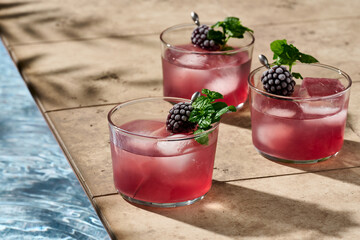 Three bramble cocktails garnished with a frozen blackberry and mint in hard light next to a pool.