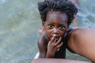 Imagen cenital de un pequeño niño afroamericano de cabello afro mirando arriba en la playa en medio de las piernas de su madre mientras mete su dedo en su boca.  