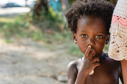 Retrato horizontal de un peque&ntilde;o ni&ntilde;o afroamericano de cabello afro al aire libre mirando a c&aacute;mara con su dedo en la boca. 