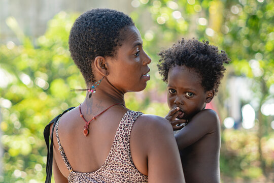 Imagen horizontal de una mujer africana de espalda cargando a su peque&ntilde;o hijo al aire libre en un d&iacute;a de verano. 