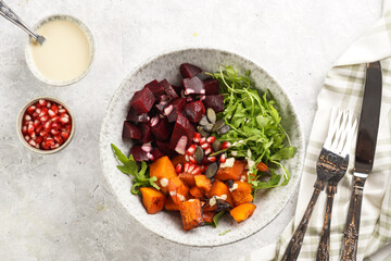 A grey deep bowl with warm winter autumn salad with arugula, red beetroot, baked pumpkin and hummus dressing cubes on a grey surface, top view