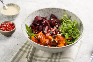 A grey deep bowl with warm winter autumn salad with arugula, red beetroot, baked pumpkin and hummus dressing cubes on a grey background
