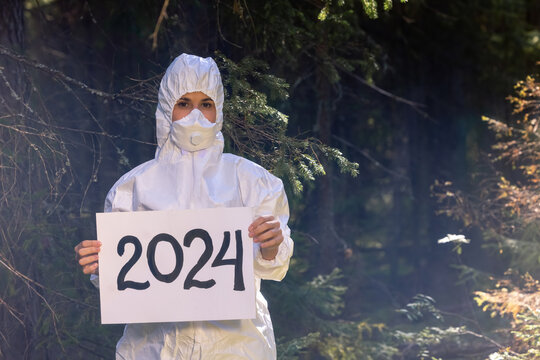 NEW YEAR 2024 - white paper plate with the inscription - 2024 year. Doctor in a white anti-epidemic suit holding a cardboard banner in hands. In the woods. 