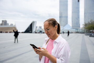 an Asian woman with glasses looks at her phone and smiles in the business district of the city