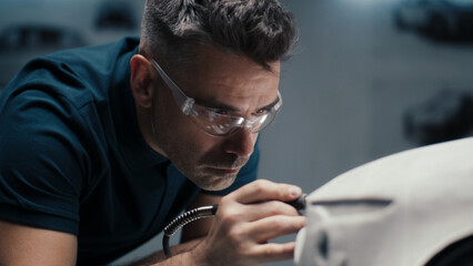 Senior automotive design engineer uses a rotary tool for perfecting the rake sculpture of a car model in a high tech company. Engineer wearing safety goggles works in an automotive company.
