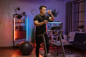 Fototapeta na wymiar Attractive young sports asian man drinking water from bottle while using smartphone, doing fitness exercise, running on treadmill . Athletic and muscular male having workout indoor at modern apartment