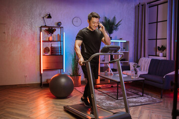 Fototapeta na wymiar Attractive young sports asian man using smartphone working out, running, doing cardio training on treadmill in evening time, indoor on background of modern apartment or gym.