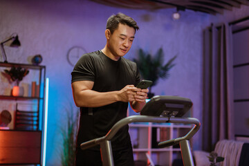 Handsome young sports asian man using phone looking online fitness class while working out, running, doing cardio training on treadmill in evening time, indoor on background of modern apartment