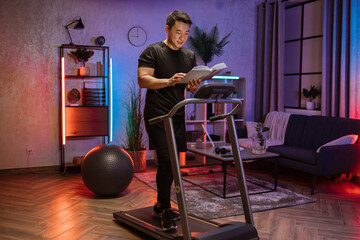 Fototapeta na wymiar Attractive young sports asian man reading book working out, running, doing cardio training on treadmill in evening time, indoor on background of modern apartment or gym.