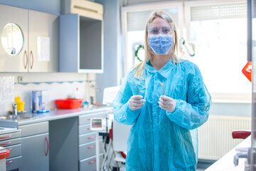 Fototapeta na wymiar Young female dentist wearing protective clothes, mask on face and face shield, protective gloves holding dental tools standing in office