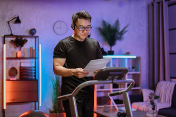 Fototapeta na wymiar Attractive young sports asian business man in headset working with papers while working out, running, doing cardio training on treadmill in evening time, indoor at home.