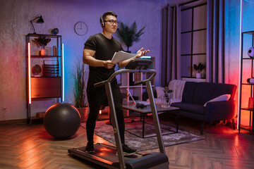 Portrait of active and dynamic young handsome businessman with headset and papers doing sport fitness at home running on treadmill indoor at night. Tone your body. Perfect shape. Improving endurance