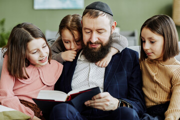 Portrait of orthodox Jewish man wearing kippah while reading book to three children - obrazy, fototapety, plakaty