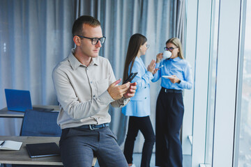 Smiling young male manager in glasses talking on the phone while standing in the office with colleagues in the background. A working day in a modern office with large windows