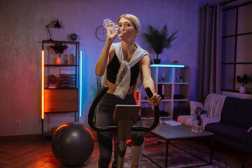 Portrait of young female having conversation on her smartphone, drinking water while working out using exercise bike. Home fitness workout sporty woman training on smart stationary bike indoors.