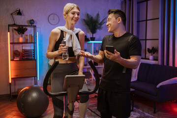 Portrait of female wearing sport bra holding bottle water working out using exercise bike and speak to asian man trainer. Home fitness workout two people training on smart stationary bike indoors.