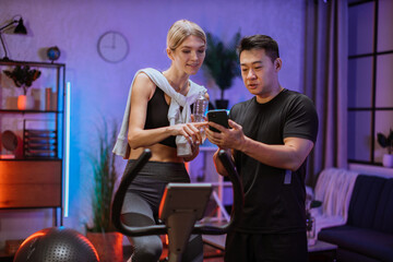 Fototapeta na wymiar Portrait of female wearing sport bra holding bottle water working out using exercise bike and speak to asian man trainer. Home fitness workout two people training on smart stationary bike indoors.
