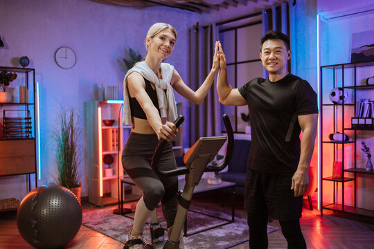 Beautiful smiling young woman doing cardio on stationary bike giving high five to her fitness trainer for good results. Attractive female working out on fitness at evening time at home .