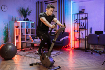 Home fitness workout, young asian man athlete training on smart stationary bike indoors watching on...