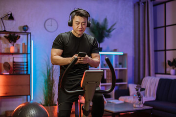 Healthy fit smiling asian man training at home on exercise static bike during workout holding phone, listening music with headphones for motivation. Male healthy weekly habits app.