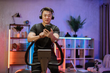Young asian man with headphones doing exercises on stationary bicycle at home or fitness center. Young sporty male in gym listen music from smartphone. Man doing cardio exercises.