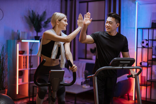 Joyful caucasian woman training on exercise bike with sporty asian man running on treadmill and giving high five. Smiling young couple doing cardio training on exercise machines at evening time